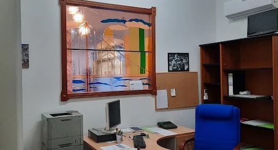 Ufficio con vetrata dipinta in cornice di legno, scrivania, computer, stampante, poltroncina blu e bacheca di sughero
